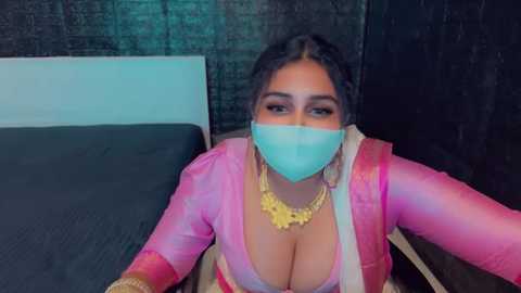 Media: Video of a South Asian woman with medium-dark skin, wearing a pink satin sari with a plunging neckline, large gold necklace, and a turquoise face mask. Background features a dark, textured wall and a black bedspread.