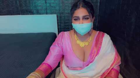 Media: Video of a South Asian woman in a pink and white sari, gold necklace, and light blue face mask, sitting on a bed with dark headboard, dark walls.