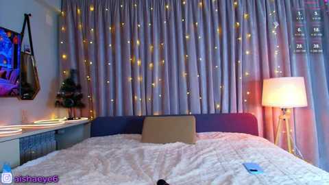 Media: Video of a modern bedroom with a soft, purple-toned bedspread, fairy lights, a blue lamp, and a laptop on the bed.