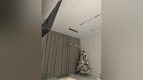 Media: Video of a minimalist room with a large gray curtain, a mounted flat-screen TV, a Christmas tree with white and red ornaments, and a coat rack in the background.