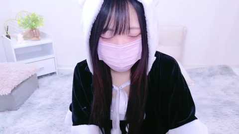 Media: Video of an Asian woman in a black and white maid costume with a pink mask, kneeling on a soft, light gray carpet in a minimalist bedroom with white walls and a white bed.