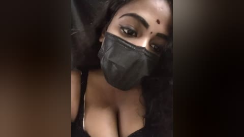 Media: A close-up video of a South Asian woman with medium-dark skin, wearing a black mask, red bindi, and black lace bra, her dark hair slightly disheveled, against a dark, blurred background.