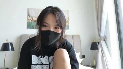 Media: Video of an Asian woman with long dark hair and a black mask, wearing a black sweatshirt with a graphic, sitting on a bed with beige headboard and white pillows.