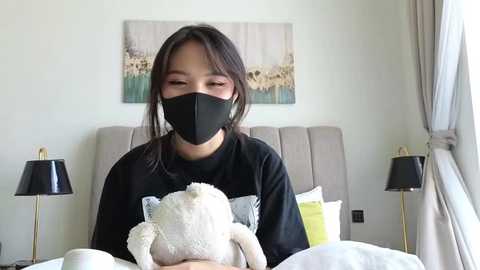 Media: A video of an Asian woman with shoulder-length black hair, wearing a black face mask and black shirt, holding a white teddy bear in a modern, minimalist bedroom with beige walls, a gray headboard, and two black table lamps.