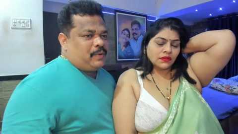 Media: Video of an Indian couple in casual attire; man in blue shirt, woman in green saree with white blouse, both smiling. Background features framed family photos, blue lighting, and a bed.