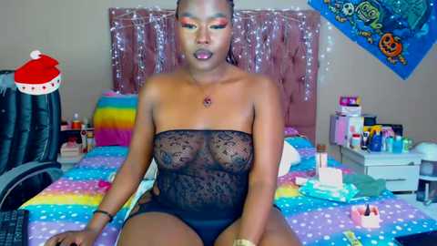 Media: Video of a dark-skinned woman with braided hair, wearing a sheer black lace bodysuit, sitting at a desk in a colorful, cluttered room with a rainbow bedspread, surrounded by makeup and jewelry.