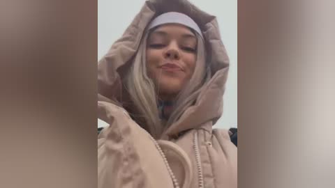 Media: Video of a blonde woman with fair skin, wearing a beige puffer jacket, white beanie, and a light blue shirt, smiling and stretching her arms above her head, indoors.