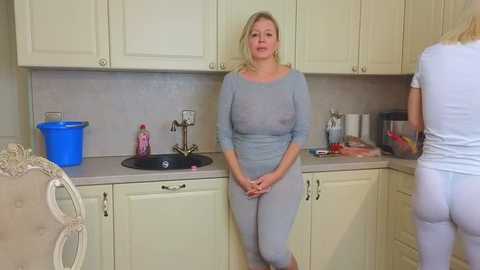 Media: A video of a middle-aged blonde woman in a light blue long-sleeve top and matching leggings standing in a cream-colored kitchen, with a woman in a white outfit visible in the background.