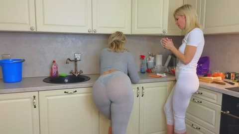 Media: Video of two blonde women in tight, light gray leggings, one washing dishes, the other standing beside her in a kitchen with white cabinets and a gray countertop.