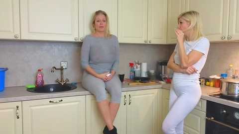 Media: Video of two blonde women in matching light grey sweatsuits sitting on a kitchen counter, discussing in a modern kitchen with white cabinets and beige countertops.