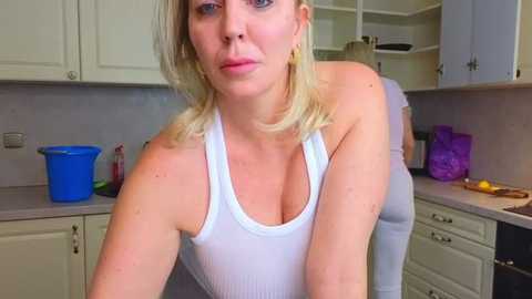 Media: Video of a blonde, fair-skinned woman in a white tank top, leaning over a kitchen counter, with a blue bucket and purple cups in the background.