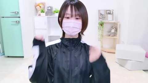 Media: Video of a woman with short, dark hair and a white surgical mask, wearing a black plastic apron, in a clean, modern room with white walls and light wooden furniture.