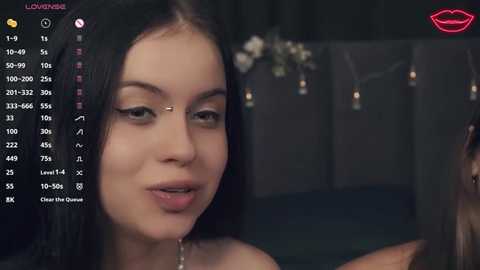 Media: Video of a close-up of a young woman with long black hair, wearing a gold nose ring, and a subtle makeup look. Background shows a dimly lit room with a bed and hanging lights.