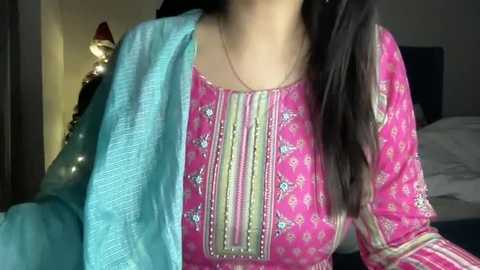 Media: Video of a woman with long dark hair wearing a bright pink salwar kameez adorned with intricate embroidery, with a turquoise dupatta draped over her shoulder. The background features a dimly lit bedroom with a bedside lamp.