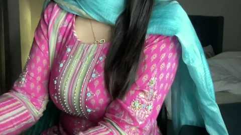 Media: Video of a woman with medium skin tone, wearing a vibrant pink traditional Pakistani dress adorned with intricate embroidery and floral patterns. She has long, dark hair and a teal scarf draped around her neck. The background features a dimly lit bedroom with a bed.