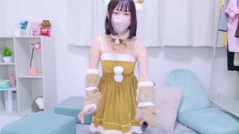 Media: Video of an Asian woman with pale skin, wearing a yellow dress with fluffy sleeves, white face mask, and bunny ears, posing in a room with pastel furniture and white walls.