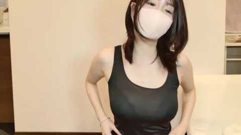 Media: A video of an East Asian woman with straight black hair, wearing a white face mask, black tank top, and white bracelet, indoors against a beige wall.