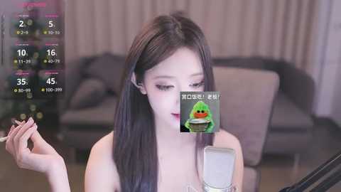 Media: A video of a young Asian woman with long black hair, wearing a white top, applying makeup in a modern living room with gray furniture. A virtual filter with a green frog emoji overlays her face.