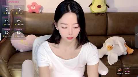 Media: Video of a young Asian woman with straight black hair, wearing a white T-shirt, sitting on a brown leather couch with plush toys and a video game screen overlay.