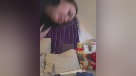 Media: A blurred video shows a person in a purple dress bending over a beige couch, surrounded by stuffed animals and a TV remote. The background is a light beige wall.