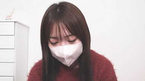 Media: Video of a young Asian woman with long, straight brown hair and bangs, wearing a white face mask and a maroon sweater, leaning forward, eyes closed, against a white background.