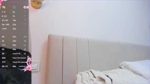 Media: Video of a modern, minimalist bedroom with a beige upholstered headboard and a neatly made bed with white sheets and a light blanket. A smart home panel with a pink flower overlay is mounted on the wall.