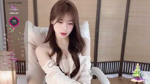 Video of an Asian woman with long brown hair, fair skin, and a slender build, sitting in a beige chair, wearing a white off-shoulder top, in a modern room with wooden partitions.