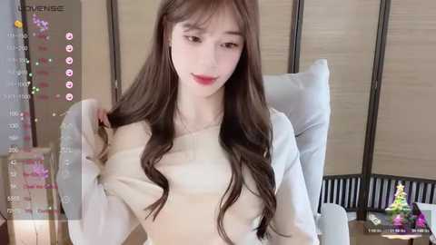 Media: Video of an East Asian woman with long, wavy brown hair and fair skin, wearing a white sweater, sitting on a white leather chair in a modern living room with beige folding screens.