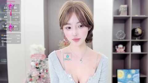 Media: Video of an East Asian woman with light skin, brown hair, and a slim figure, wearing a light grey cardigan and a pendant necklace. She stands in a modern room with bookshelves and decorative items in the background.