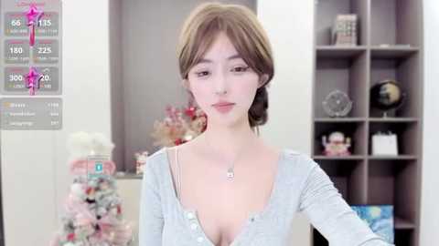 Media: Video of a young woman with light skin and brown hair, wearing a low-cut, light gray cardigan. Background features a white room with a pink tree, a globe, and a bookshelf.