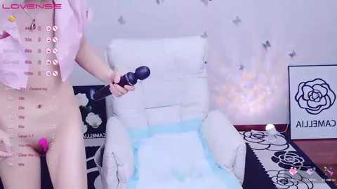 Media: Video of a light-skinned person in a pastel pink robe, holding a black vibrator, standing in a room with white furniture and a black floral-patterned rug.