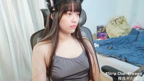 Media: A young Asian woman with long brown hair and black cat ears, wearing a grey tank top, sits on a black office chair in a messy bedroom with a bed and blue sheets.