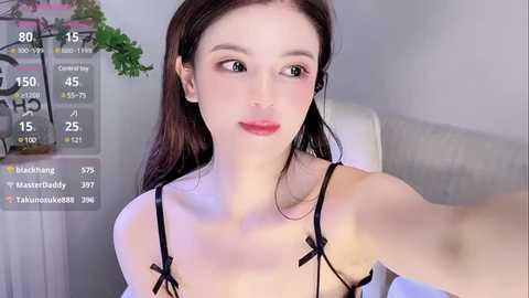 Media: A video of a pale-skinned, petite Asian woman with long brown hair, wearing a black lingerie top with ribbon straps, sitting in a modern living room with a white couch and green plants.