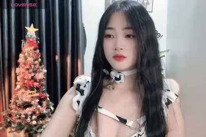 Media: Video of an East Asian woman with long black hair, wearing a revealing white and black cow-print lingerie set, standing in front of a decorated Christmas tree and black curtains.