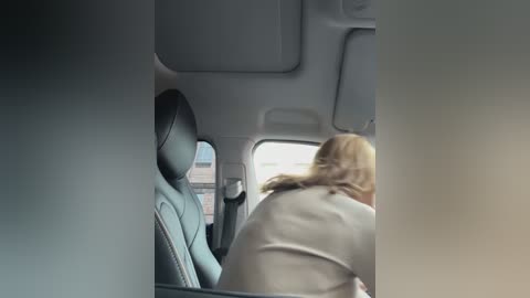 Media: Video of a passenger in a moving car, viewed from behind. The person, with shoulder-length blonde hair, wears a beige coat. The car's interior features gray leather seats and a tinted window showing an urban landscape.