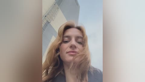 Media: Video of a young, fair-skinned woman with long, wavy, light brown hair, wearing a black jacket, standing between two vertical columns against a cloudy sky background.