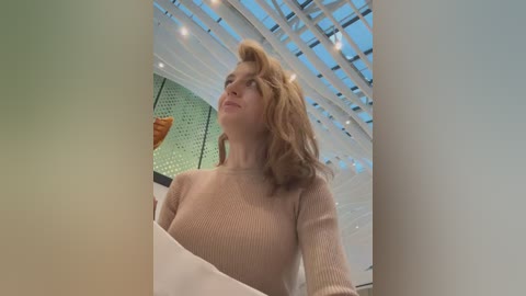 Media: Video of a fair-skinned woman with wavy, shoulder-length blonde hair, wearing a beige sweater, standing indoors with a modern, white, grid-patterned ceiling. Background includes a partially visible orange and green artwork.