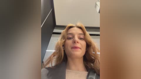 Media: Video of a young woman with wavy, shoulder-length blonde hair, wearing a light sweater and gray jacket, standing in a modern, minimalist room with beige walls and recessed lighting.