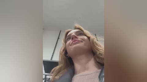 Media: Video of a smiling, fair-skinned woman with blonde, wavy hair, wearing a light pink top and a gray cardigan, looking upwards with a slight tilt. The background features a white ceiling and part of a kitchen.