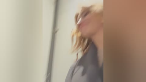 Media: A blurry video of a person with light skin and wavy blonde hair, wearing a grey jacket, standing against a beige wall, with a blurred background.