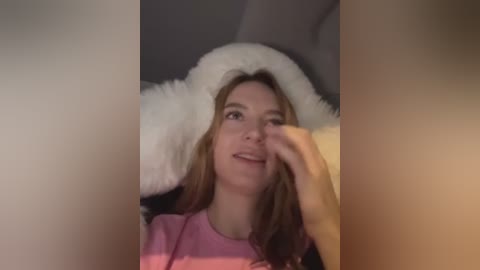 Media: Video of a young Caucasian woman with light skin and long brown hair, wearing a pink t-shirt, holding a fluffy white teddy bear, smiling, in a dimly lit room.