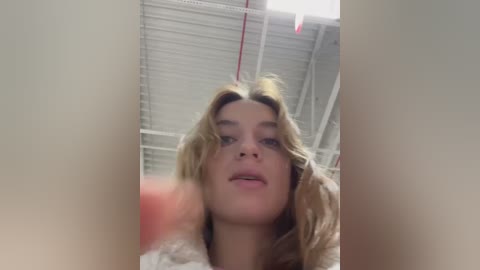 Media: Video of a young Caucasian woman with wavy blonde hair, wearing a beige jacket, taken indoors with a white ceiling and red cords visible.