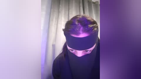 Media: A video of a person with a black blindfold, wearing a black shirt, standing in front of a grey curtain, bathed in soft purple light.