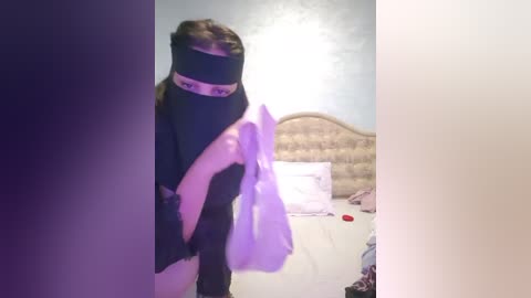 Media: Video of a woman wearing a black blindfold, purple mask, and black clothing, standing in a bedroom with a white bed, a wicker headboard, and scattered clothes.