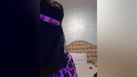 Media: Video of a woman in a black hijab with a purple pattern, standing in a bedroom with a light blue textured wall, a patterned headboard, and white bed linens.