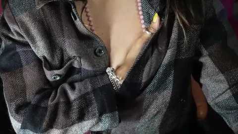 Media: Close-up video of a woman's partially unbuttoned, grey plaid shirt revealing a pink lace bra and a silver necklace with a yellow pendant.