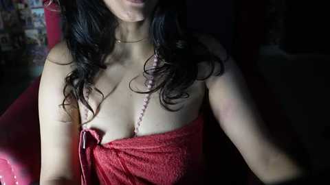 Media: Video of a woman with long, wavy black hair, wearing a red, strapless top, exposing cleavage, in a dimly lit room with a pink chair and blurred background.