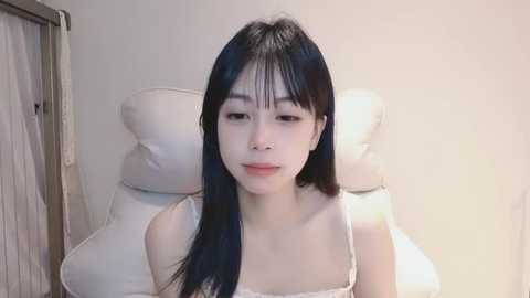 Media: A video of an Asian woman with long black hair, wearing a white spaghetti-strap top, sitting on a white cushioned chair against a plain white wall. She has a serene expression.