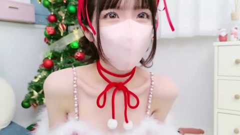 Media: Video of an Asian woman with fair skin, wearing a red ribbon choker and white face mask, in a festive room with a decorated Christmas tree and white dresser.