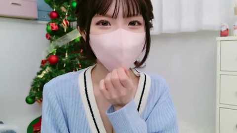 Media: Video of a young Asian woman with straight black hair, wearing a light blue sweater and a pink face mask, blowing her nose indoors near a decorated Christmas tree.